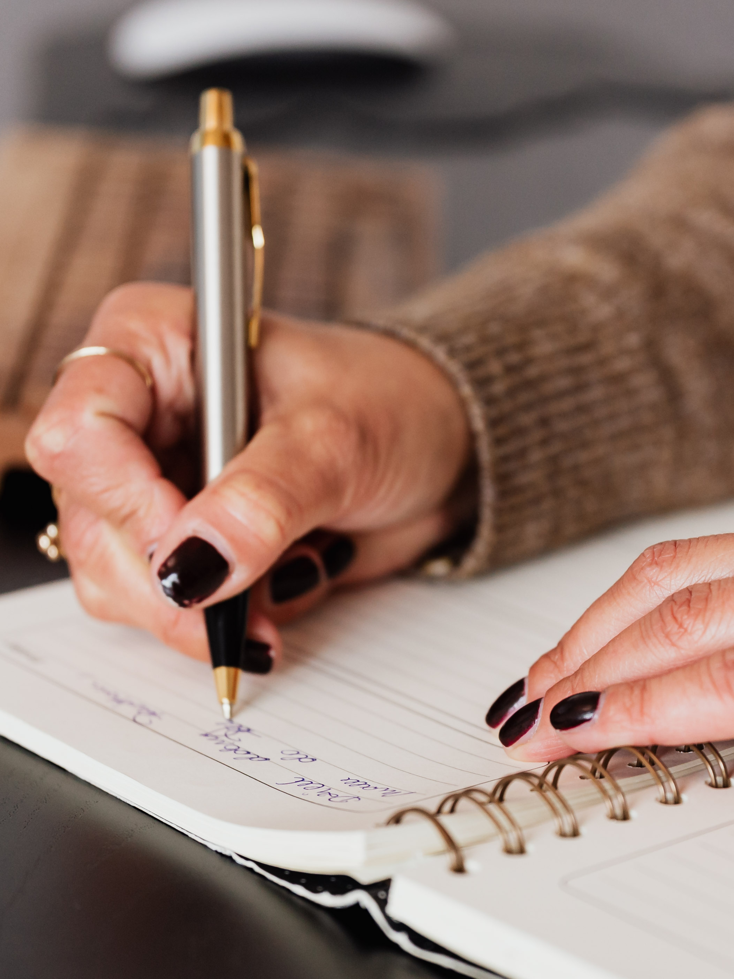 Female hands with gold rings and black painted nails holding a silver pen while writing in a notebook.